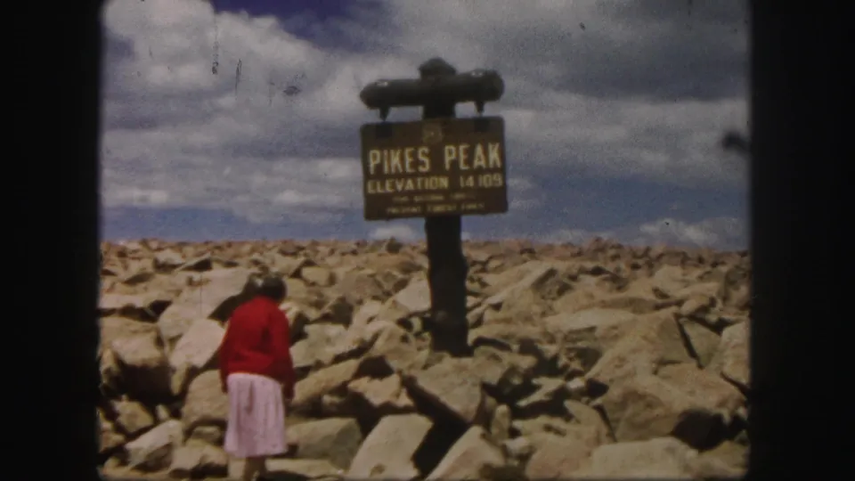The woman moves closer to the Pikes Peak sign, with boulder-strewn summit terrain and a cloudy sky visible.