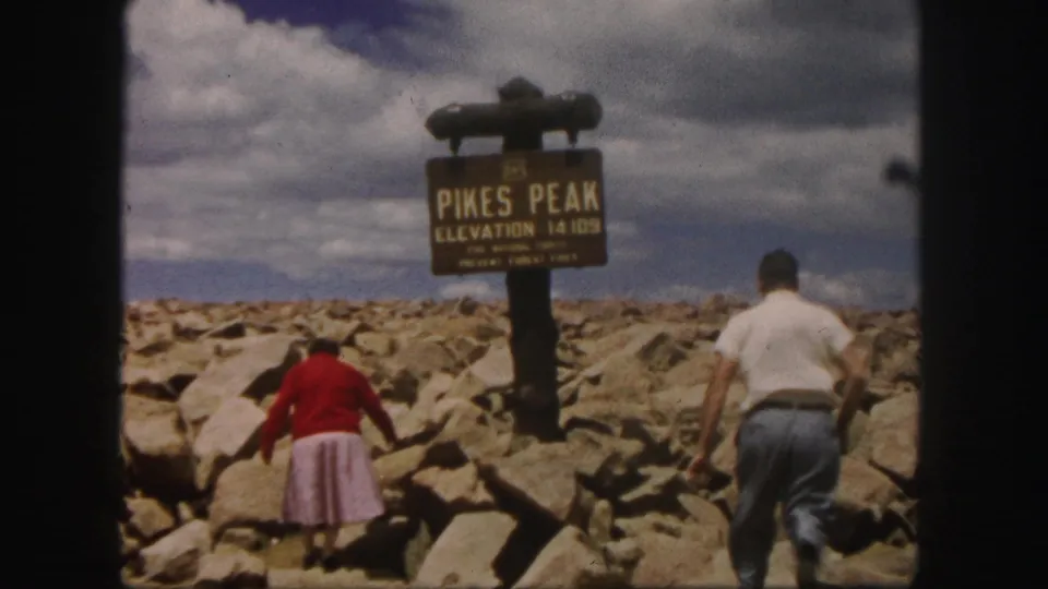 A man in a white shirt enters as the woman continues toward the Pikes Peak sign on the rocky Colorado summit.