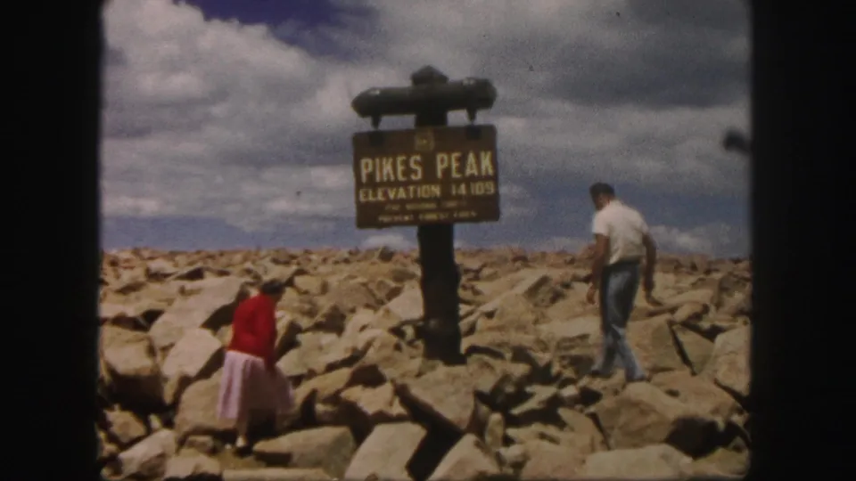 The man and woman walk separately over rocks below the Pikes Peak elevation sign, filmed in a handheld home-movie view.