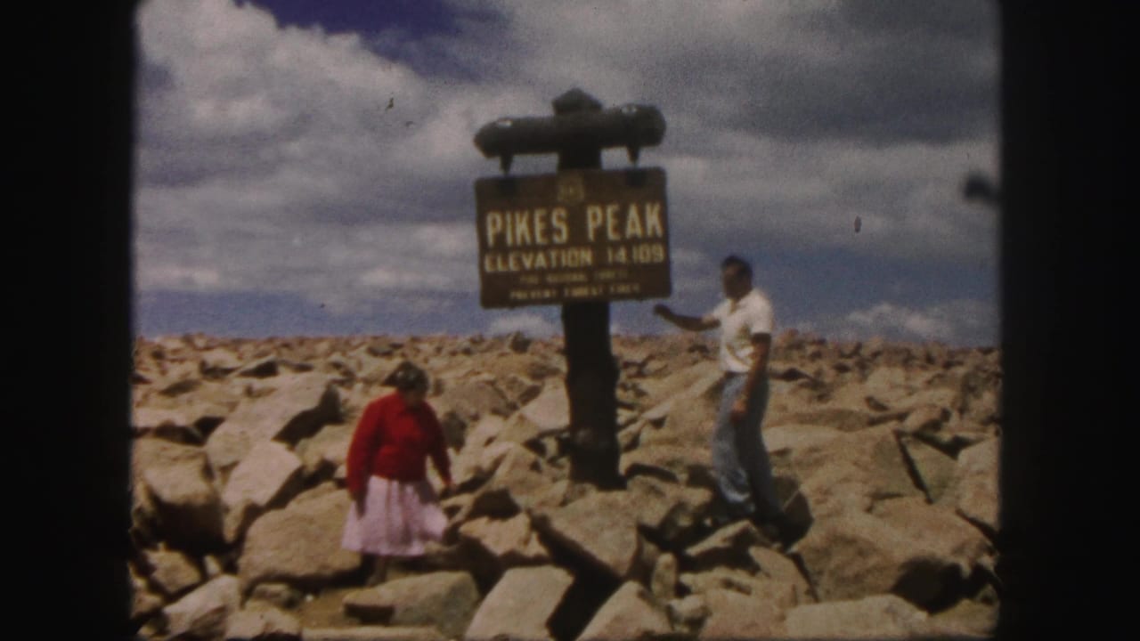 1958 Colorado home-movie frame of a man and woman posing on the rocky Pikes Peak summit beside the elevation sign.