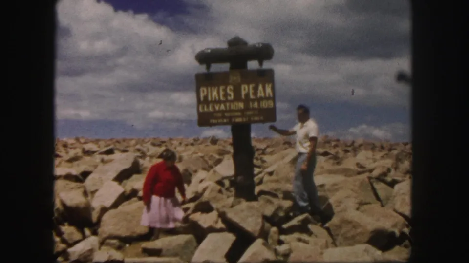 1958 Colorado home-movie frame of a man and woman posing on the rocky Pikes Peak summit beside the elevation sign.