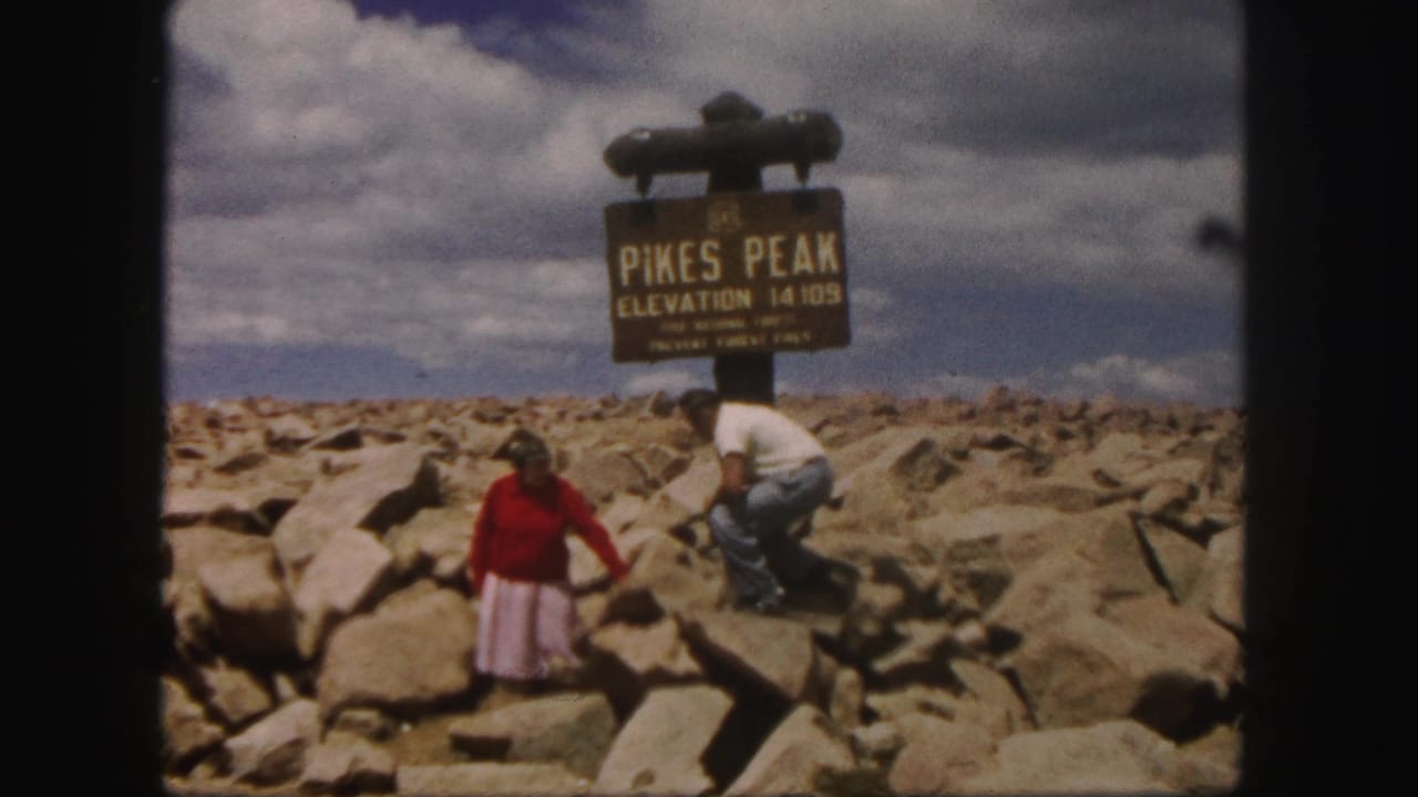 1958 Pikes Peak summit frame with a woman in red standing forward and a man crouching near the elevation sign.