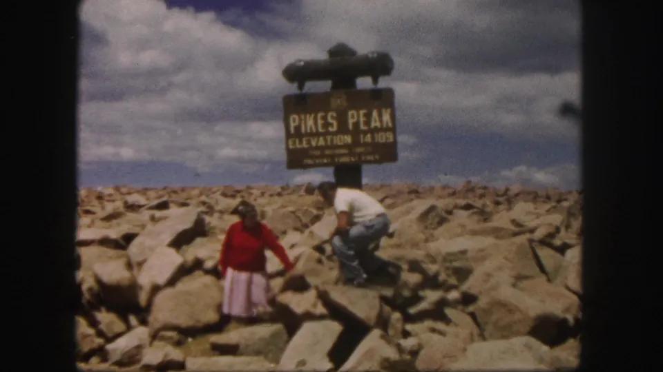 1958 Pikes Peak summit frame with a woman in red standing forward and a man crouching near the elevation sign.