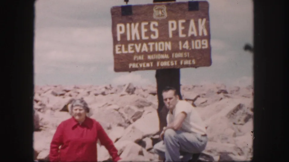 Closer 1958 home-movie view of two adults posing below the Pikes Peak elevation sign on the rocky summit.