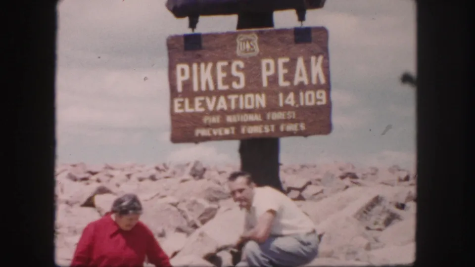 1958 Colorado Springs travel frame showing two adults below the Pikes Peak summit sign, with rocky ground and pale sky behind them.