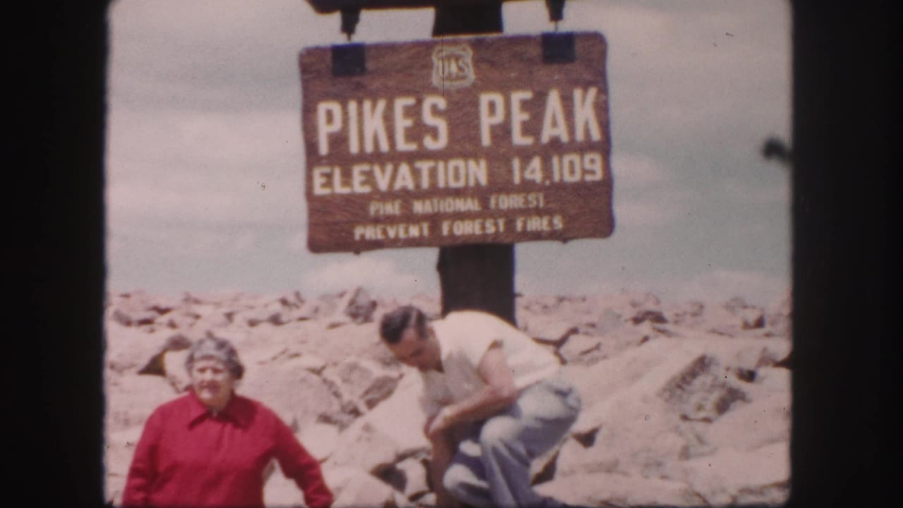 1958 Colorado home-movie frame showing two adults near the Pikes Peak elevation 14,109 sign on rocky summit ground.