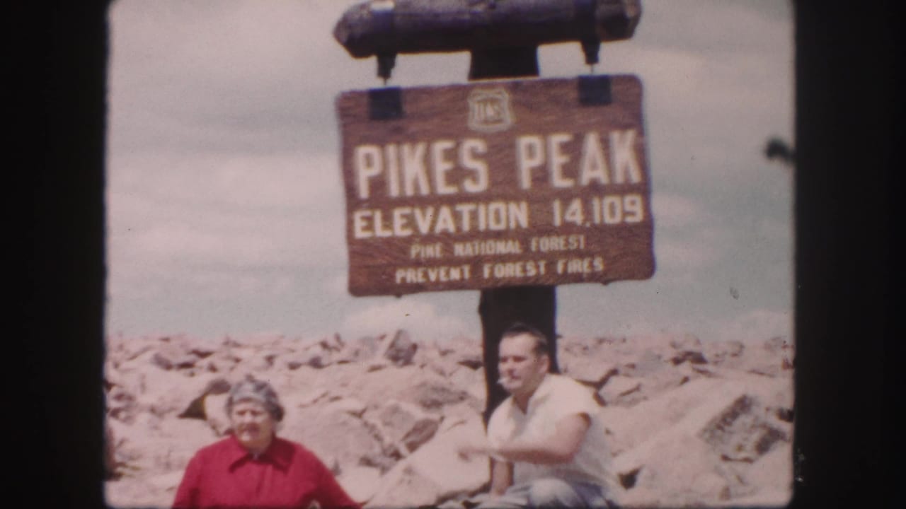 A woman and man sit or crouch by the Pikes Peak elevation sign.