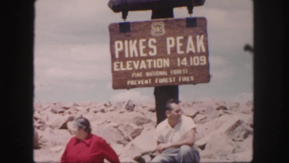 1958 home-movie frame of two adult visitors seated below the Pikes Peak sign, surrounded by pale rocky summit terrain.