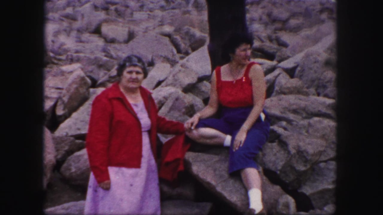 In Colorado Springs, 1958, two adult women pose on a rocky outcrop, one standing in a red jacket and light dress and the other seated in red and blue attire.