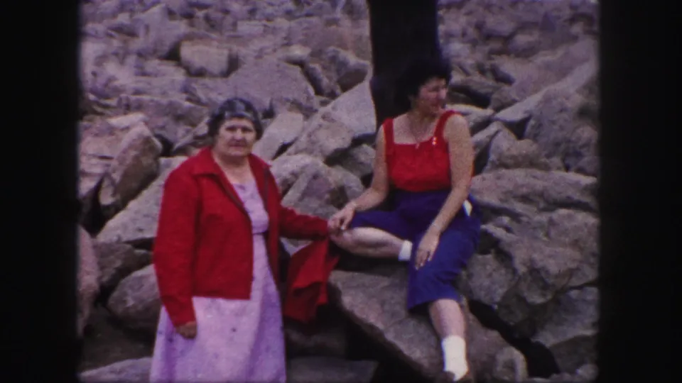 In Colorado Springs, 1958, two adult women pose on a rocky outcrop, one standing in a red jacket and light dress and the other seated in red and blue attire.