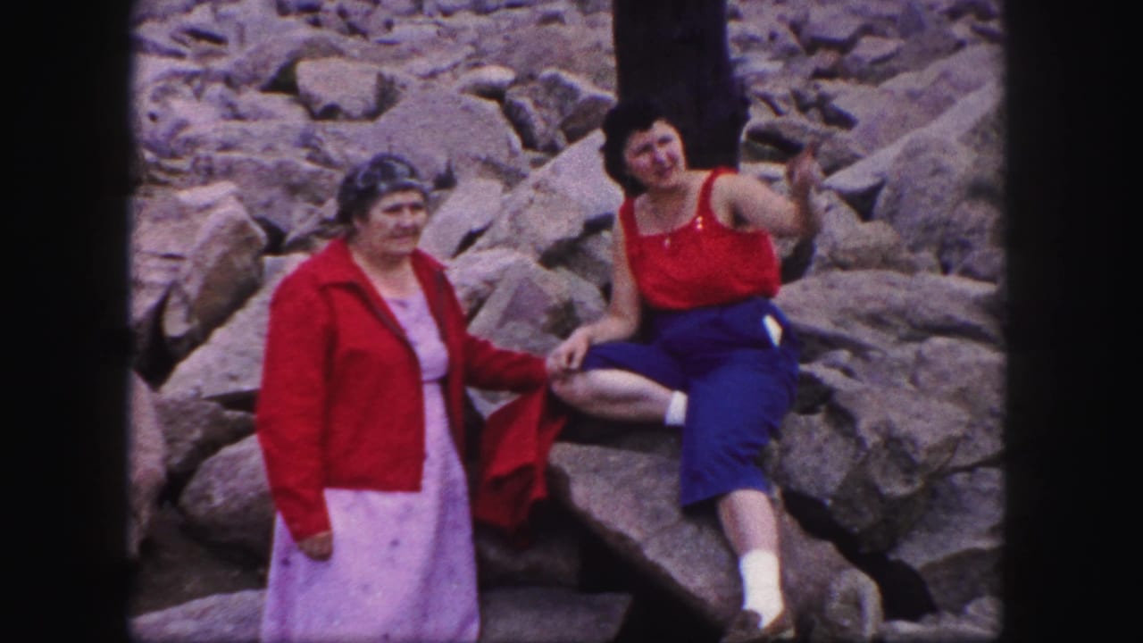 One woman stands on rocks while another sits beside her and points outward from the rocky setting.