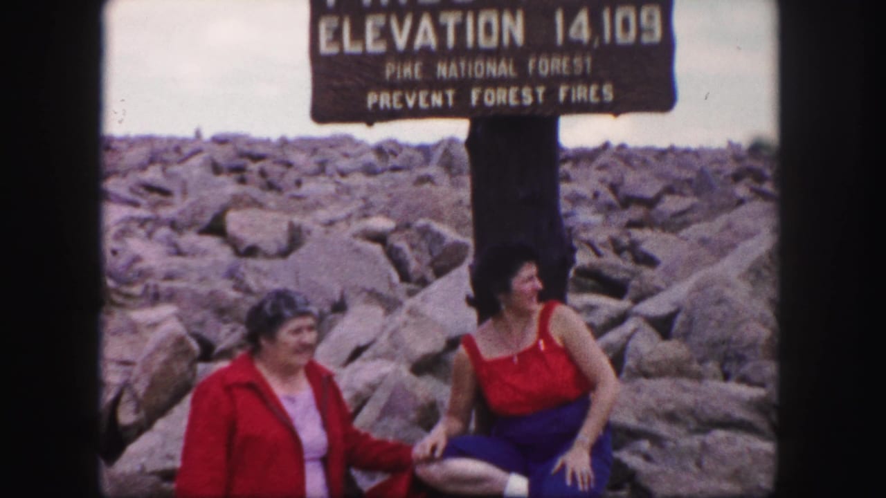 Two women sit and stand beneath the Pikes Peak elevation sign.