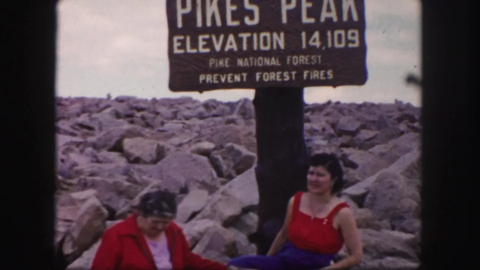 The Pikes Peak elevation sign is prominent above two women seated and standing on summit rocks.