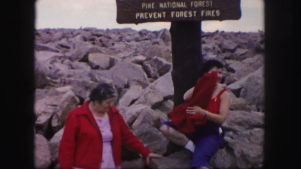 Two women rest near the Pikes Peak summit sign while one raises a red jacket or garment.