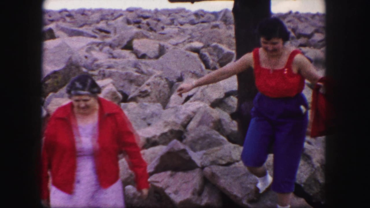 Two women step carefully across large rocks in an outdoor area.