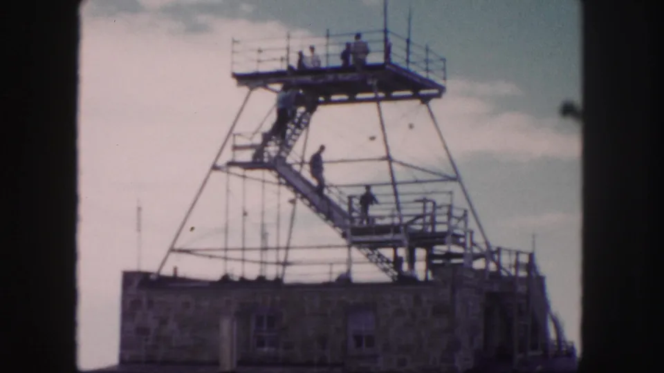 1958 Colorado Springs home-movie frame showing small visitor figures on a metal stairway and platform above a stone building.