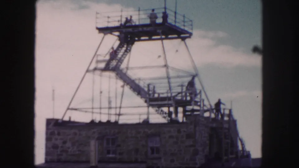 Final 1958 Colorado Springs tower frame showing visitors dispersed on the stairway and elevated platform over a stone structure.