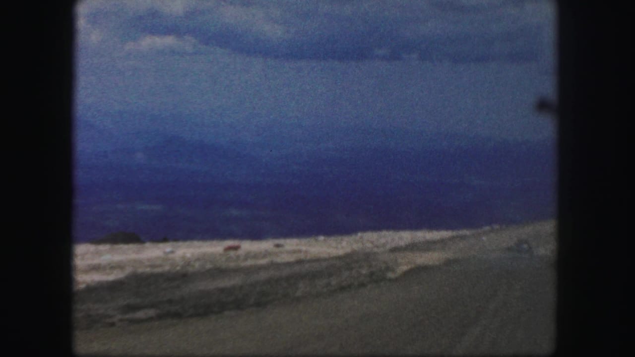 1958 Colorado Springs 8mm frame from a moving vehicle, looking over roadside terrain toward a hazy valley and dark grey clouds.