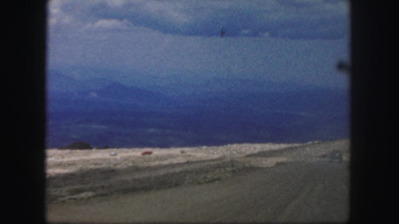 Open valley and distant blue mountains seen beyond a rough roadside under low grey clouds.