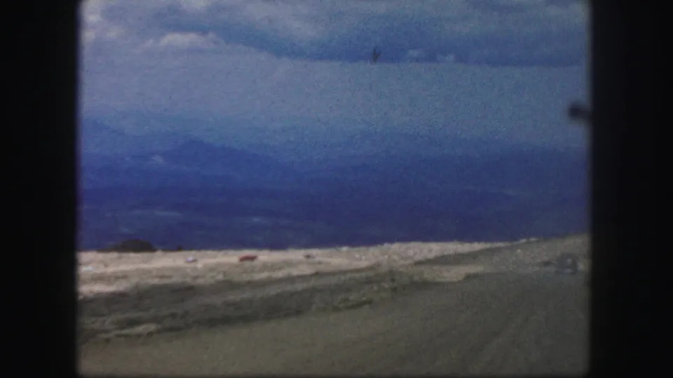 Vehicle-shot 1958 Colorado Springs view with rocky roadside, broad valley distance, blue mountain haze, and low grey sky.