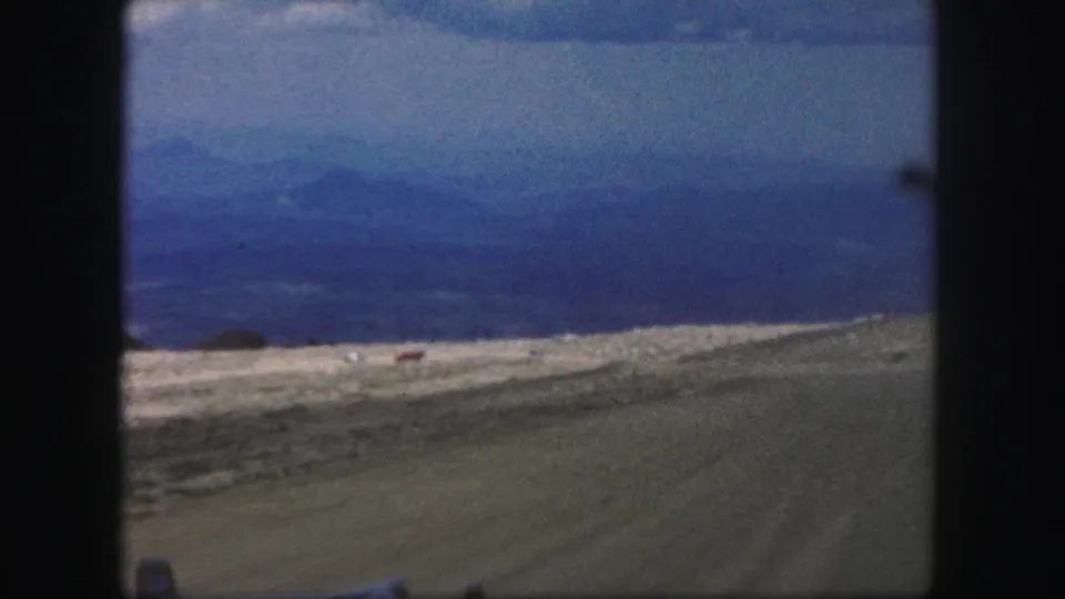 Final 1958 Colorado Springs frame showing a dirt foreground and open valley under hazy blue mountains and a grey sky, framed by 8mm borders.