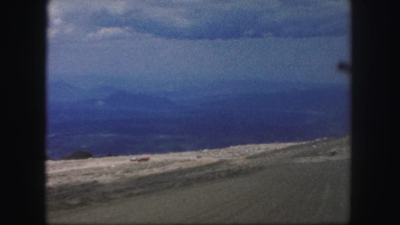 Colorado Springs-area mountain view from 1958 8mm footage, with a small red vehicle on a pale road across open hills.
