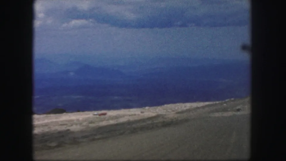 Colorado Springs-area mountain view from 1958 8mm footage, with a small red vehicle on a pale road across open hills.