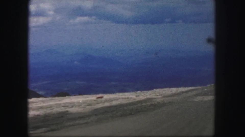 1958 home-movie frame near Colorado Springs showing layered blue mountains beyond a sparse hillside and roadside strip.