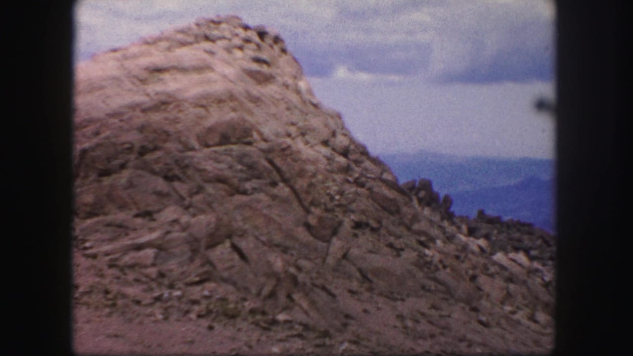 1958 Colorado Springs home-movie frame showing a rugged rock formation, gravel slope, cloudy sky, and distant blue hills.
