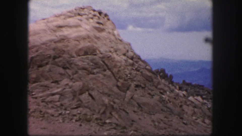 1958 Colorado Springs home-movie frame showing a rugged rock formation, gravel slope, cloudy sky, and distant blue hills.