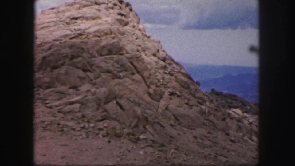 1958 Colorado Springs frame with a close view of layered rock, loose stones, muted sky, and distant mountain ridges.