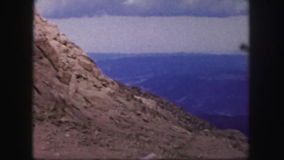 1958 Colorado Springs frame showing the edge of a rock formation, sloping terrain, distant valley floor, and layered blue ridges.