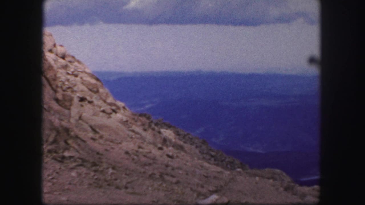 1958 8mm Colorado Springs home-movie frame showing a sunlit rocky slope above a broad blue distant landscape under clouds.