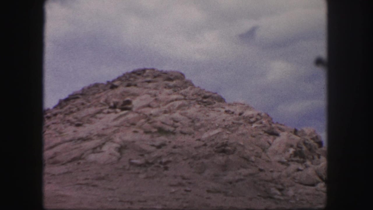 Rocky mound seen through a dark vehicle window frame under a cloudy sky.