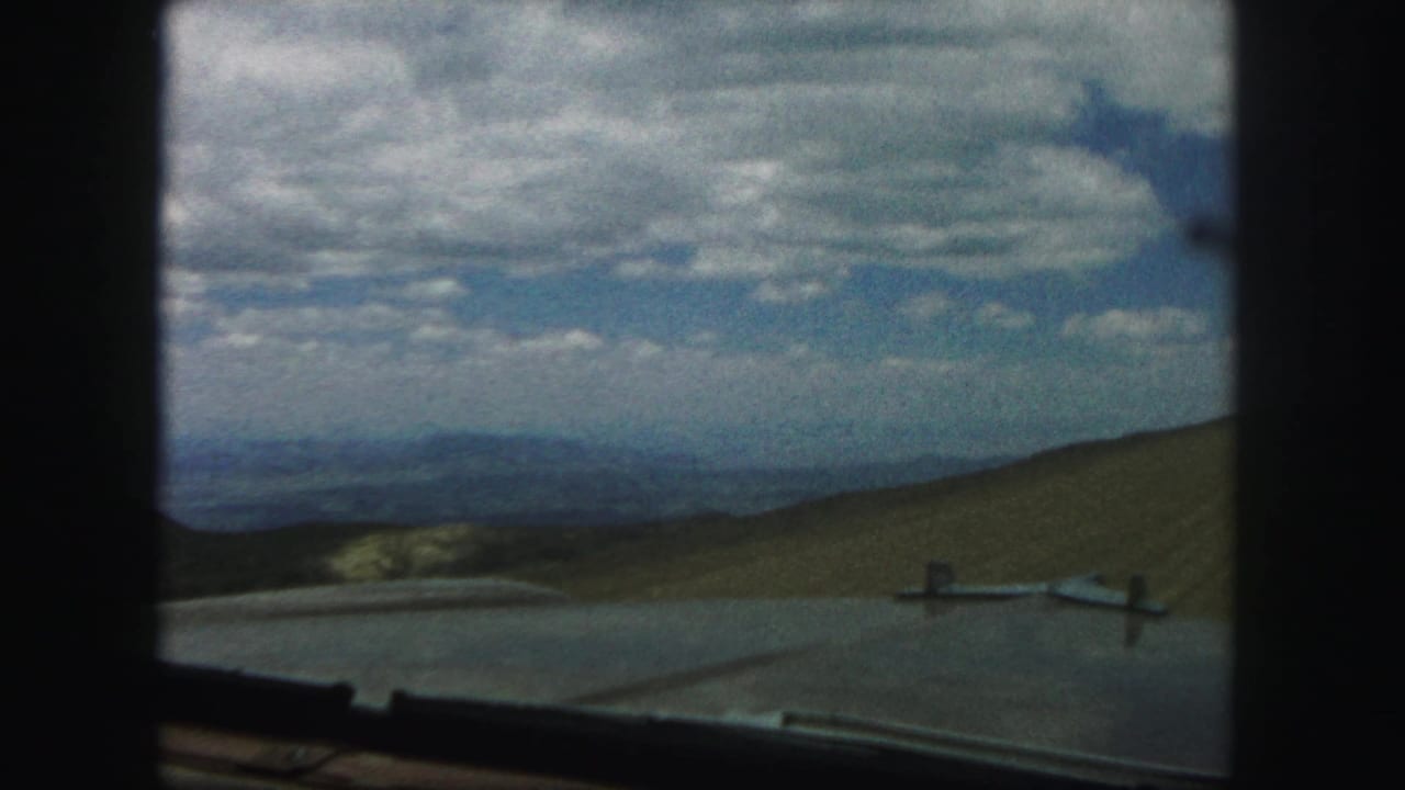 View through a car window toward grassy hills and a cloud-filled blue sky.