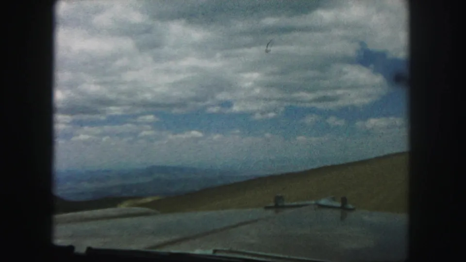 1958 home-movie road view showing a sloped grassy hillside, distant blue terrain, and textured cloud cover through a car window.