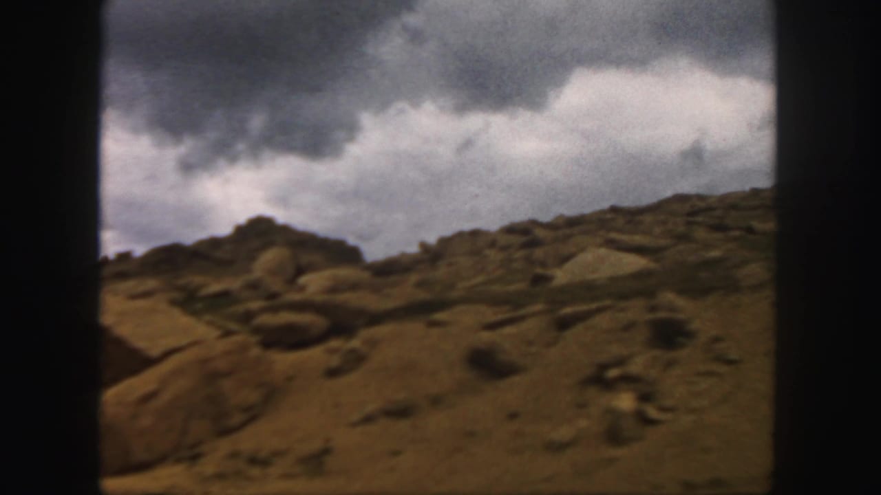 1958 Colorado 8mm view of a rocky mountain slope under heavy grey and white clouds, framed by dark film edges.