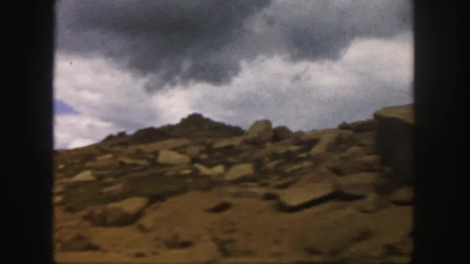 1958 Colorado 8mm frame showing a rough boulder-covered ridge below dark clouds and a small break of blue sky.