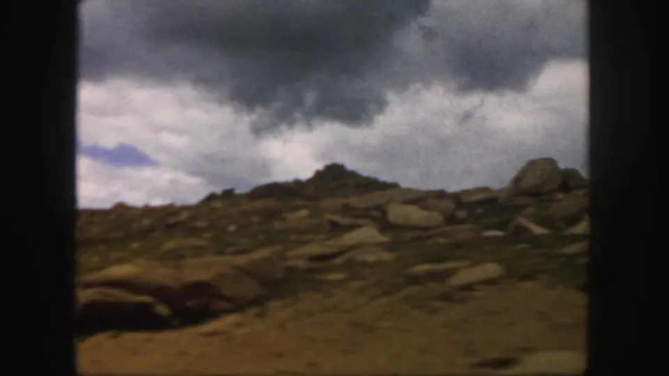 1958 Colorado 8mm view across uneven rocky terrain toward a low ridge under grey clouds and a narrow blue opening.