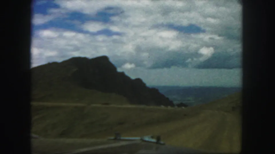 1958 Colorado 8mm frame with a winding road crossing open hills toward a dark mountain, cloudy sky, and distant blue horizon.