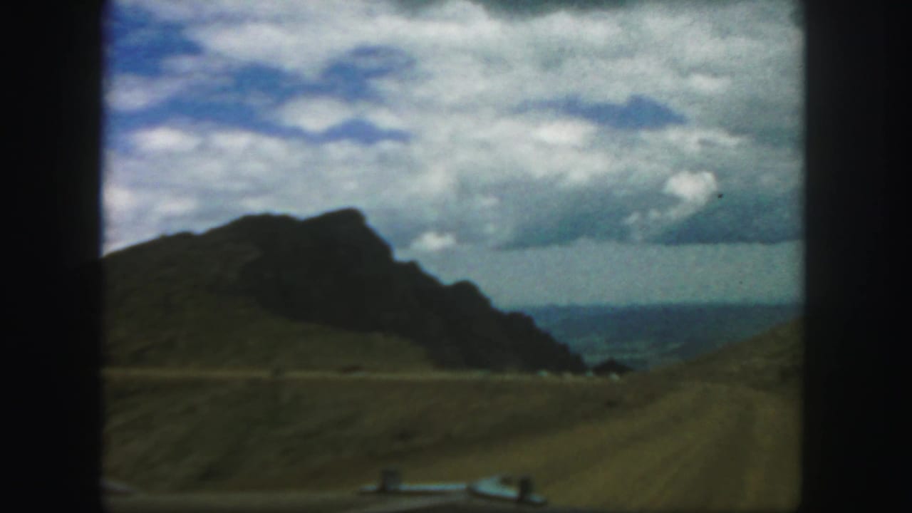 1958 Colorado 8mm frame from a moving car, showing a dirt mountain road, grassy slopes, rocky hill, and cloudy sky.