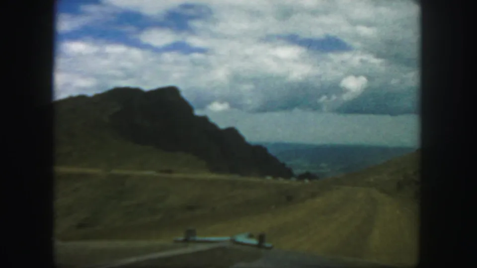 1958 Colorado road-trip view from inside a car, with a winding dirt road crossing open hills toward rugged mountains.