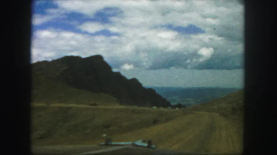 1958 Colorado 8mm travel frame showing a dirt road through grassy mountain terrain with distant blue water or haze beyond.