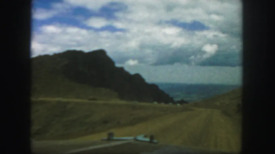 1958 Colorado home-movie frame from a car, looking along a rural dirt road between grassy hills and a rugged mountain form.