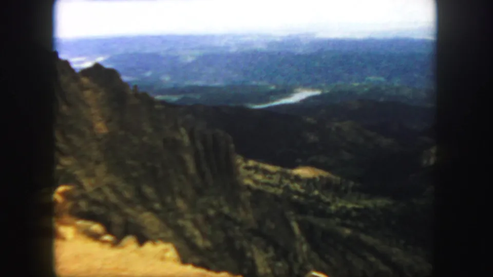 1958 Colorado home-movie frame with rugged foreground rock, forested slopes, distant ridges, and a pale river curve in the valley.