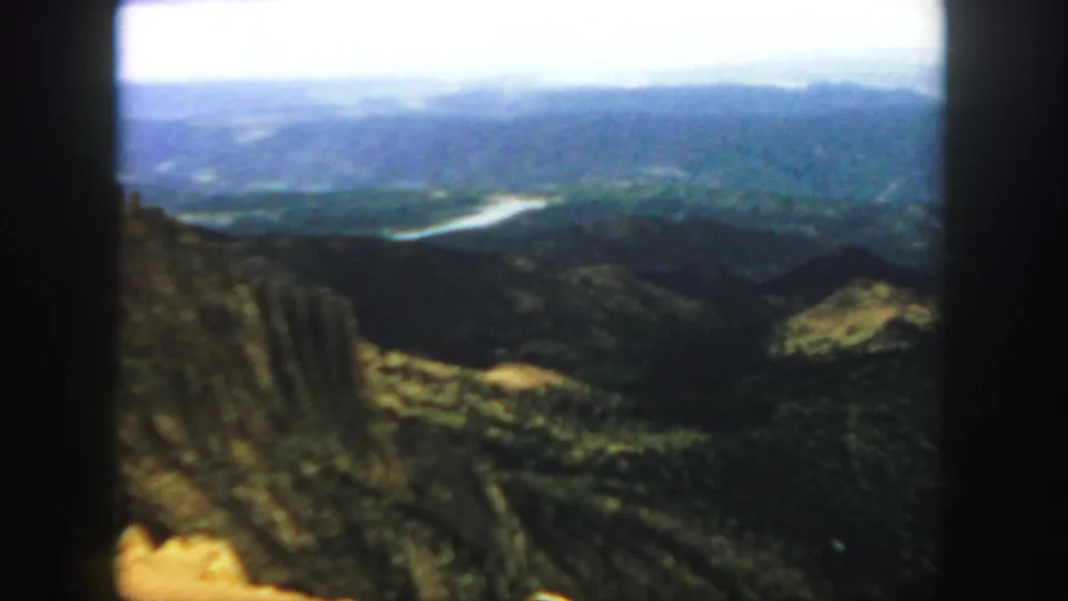 1958 Colorado home-movie frame with a wide mountain landscape, rocky ridge in front, green slopes, and a pale river in the distance.