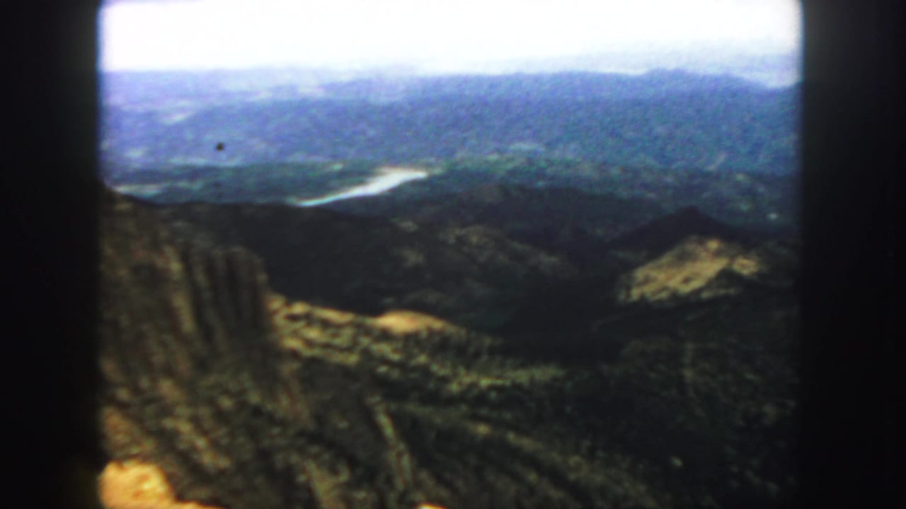 Soft 8mm view over mountain ridges, wooded slopes, and a pale winding river below.