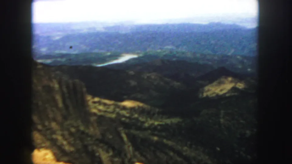 1958 Colorado 8mm frame showing a high view across layered mountains, dark valleys, and a bright river or reservoir in the distance.