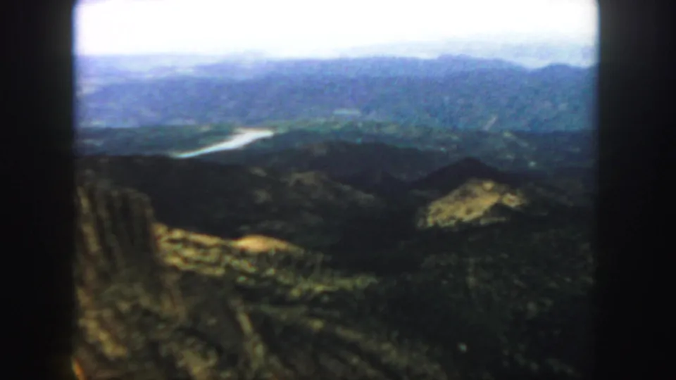 1958 Colorado home-movie frame with layered ridges, green-brown slopes, and a light-toned river or reservoir cutting through the valley.