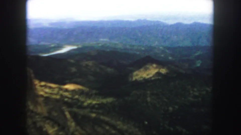 1958 Colorado 8mm still showing a broad hazy mountain panorama, foreground hills, and a pale winding river or reservoir in the valley.